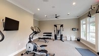 Located at 18172 Section St in Fairhope, Alabama, this dedicated fitness center offers a thoughtfully designed, single-floor workout space suited for a comprehensive exercise routine. The room features neutral, warm-toned walls accented by crisp crown molding, recessed lighting, and a ceiling fan that keeps the environment comfortable during workouts. Plush, low-pile carpeting covers the floor throughout, providing a clean and cushioned surface underfoot.

The space is well-appointed with a diverse array of fitness equipment. Cardio options include multiple elliptical trainers, a treadmill, and what appears to be a rowing machine, all positioned to take advantage of wall-mounted flat-screen televisions. For strength training, the room is equipped with a dual-cable functional trainer, a multi-tiered dumbbell rack stocked with a full range of free weights, kettlebells, and an adjustable weight bench.

Large windows along one wall flood the interior with natural light and offer views of the surrounding greenery, creating an inviting and energizing atmosphere. Additional amenities include wall-mounted displays for monitoring workouts and organized storage baskets for towels and accessories. The overall design strikes a balance between functionality and comfort, making this fitness center a practical and well-maintained facility for daily use.