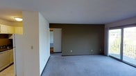 This single-floor space presents a straightforward, functional layout with a clean and understated aesthetic. The main room is finished with wall-to-wall grey carpeting, providing a soft, neutral foundation that complements the surrounding color palette. Two distinct wall treatments define the room: crisp white walls on one side contrast with a deep taupe-brown accent wall, adding visual depth and a subtle design element to the otherwise minimalist interior.

The ceiling is smooth and white, contributing to a bright, open feel throughout the space. Multiple electrical outlets are evenly distributed along the walls, offering practical utility for various furniture arrangements. A light switch is visible near the entry point, indicating thoughtful placement for ease of access.

An open doorway along one wall connects the carpeted main room to an adjoining area, which features hard surface flooring, suggesting a functional transition into a kitchen, hallway, or secondary room. This glimpse into the connecting space hints at a multi-room layout within the single-floor footprint.

Overall, the space presents a clean, move-in-ready environment with a neutral design palette suited to a variety of interior styles and uses.