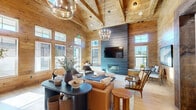 Located at 3221 NC 87 Hwy in Sanford, North Carolina, this single-floor living space showcases exceptional rustic-modern craftsmanship throughout. The open-plan living room is defined by dramatic vaulted ceilings with exposed natural timber trusses that draw the eye upward, while floor-to-ceiling tongue-and-groove wood paneling on the walls creates a warm, cohesive aesthetic. Light hardwood flooring complements the natural wood tones throughout the space.

Abundant natural light pours in through a generous arrangement of double-hung windows, including elevated transom windows that amplify the sense of height and airiness. A cozy built-in window seat invites relaxation beneath one of the window groupings. The living area is anchored by comfortable leather seating, a modern fireplace, and an eye-catching decorative feature wall with a distinctive patterned tile backdrop framing a well-appointed wet bar with a beverage refrigerator and ample counter space.

Elegant dual chandelier lighting fixtures add a refined touch to the rustic setting, while a mounted television provides a contemporary amenity. The space also features a dining nook and additional seating areas, making it highly versatile. A glass-paneled entry door connects the interior to the outdoors, reinforcing the space's bright, welcoming character throughout.