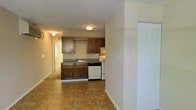 This single-floor space offers a practical and functional layout designed for ease of living. The kitchen is a central feature, outfitted with rich dark wood cabinetry that provides ample storage space both above and below the counters. A decorative tile border detail accents the upper cabinets, adding a touch of character to the space. The kitchen is equipped with built-in appliances including a white electric range, a dishwasher, and an under-cabinet range hood, making it well-suited for everyday cooking needs. A sink is conveniently positioned beneath the upper cabinetry along the main counter run. The flooring throughout the kitchen and adjoining area features durable tile in warm earth tones, providing a cohesive and easy-to-maintain surface. The open layout connects the kitchen to the adjacent living area, creating a sense of spaciousness and ease of movement. Walls are finished in a neutral palette, complemented by a textured ceiling that adds subtle visual interest. Flush-mount ceiling light fixtures provide consistent overhead illumination throughout. A set of bifold closet doors is visible, offering additional built-in storage. A hallway leads further into the unit, suggesting additional rooms beyond the main living and kitchen area.
