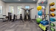 This single-floor fitness center is thoughtfully designed to accommodate a wide range of workout styles and fitness goals. The facility is organized into several distinct zones, each tailored to a specific type of training. The cardio area features multiple treadmills, elliptical trainers, and a rowing machine, all positioned along walls lined with large windows that invite generous natural light and outdoor views, creating an energizing environment for aerobic exercise. A dedicated strength and free weights zone houses a full rack of dumbbells, adjustable benches, and large wall-mounted mirrors that visually expand the space and assist with form. An additional functional training area offers a multi-cable resistance machine, a weight bench with plate storage, stability balls, resistance bands, and exercise mats, supporting a variety of conditioning and flexibility exercises. Stationary bikes and a wall-mounted flat-screen television round out the equipment offerings in a separate section of the floor. Throughout the facility, the interiors are finished with neutral gray-toned carpet tiles and crisp white walls accented by a muted blue ceiling, lending the space a clean, modern aesthetic. Flush and recessed linear lighting ensures consistent illumination across all zones, while the entry area features light hardwood-style flooring and interior windows providing sightlines into the workout spaces.