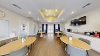Located at 3300 Winterhaven St in Las Vegas, NV, this single-floor community dining room is a thoughtfully designed gathering space that balances style and functionality. The room's most distinctive architectural feature is a recessed tray ceiling with a warm yellow interior and a bold drum pendant light, complemented by recessed lighting throughout that ensures the space is well-lit at all times. Durable wood-look flooring runs the length of the room, lending a warm and cohesive aesthetic underfoot. Multiple round dining tables with modern chairs are arranged throughout the open floor plan, creating an inviting environment for communal dining and socializing. Tall French doors along one wall flood the interior with abundant natural light and provide a direct view to the outdoors, framed by floor-to-ceiling navy blue curtains that add a rich, contrasting accent to the neutral white walls. A built-in white cabinetry refreshment station along one wall features a granite countertop, sink, and coffee maker, offering practical amenities for residents. A wall-mounted flat-screen television is positioned nearby for added convenience. A small workstation with a desk and computer is tucked near the French doors, and framed botanical artwork decorates the walls, contributing to the room's clean, contemporary ambiance.