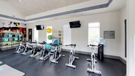 This fitness studio is housed within a single-story space distinguished by soaring vaulted ceilings with a wood-panel finish and bold exposed beams that lend the room architectural character. The generous ceiling height creates an open, airy atmosphere that enhances the sense of space throughout the studio. Dark, durable flooring runs the length of the room, providing a practical and polished surface suited for a variety of workouts.

A row of professional spinning bikes lines one side of the studio, offering dedicated space for cycling classes and individual cardio sessions. Multiple yoga and exercise mats are laid out across the open floor area, accommodating group fitness classes or solo stretching and floor work. Equipment storage racks hold an assortment of stability balls in various sizes, while a separate rack displays a graduated set of kettlebells, making strength and functional training readily accessible.

Multiple windows along the walls invite abundant natural light into the studio and provide views of the surrounding outdoor area, including a glimpse of a pool. Wall-mounted flat-screen televisions are positioned throughout for entertainment or instructional use. A bold, large-scale athletic mural serves as a dynamic focal point, contributing energy and visual interest to the overall ambiance of the space.