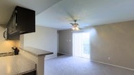 This single-floor space offers a clean, functional layout designed for comfortable everyday living. The main living area is finished with plush wall-to-wall carpeting that adds warmth underfoot, while neutral-toned walls provide a versatile backdrop for any style of furnishing. A multi-blade ceiling fan with an integrated light fixture is centrally positioned overhead, offering both ambient lighting and year-round climate comfort. A large sliding glass door dressed with vertical blinds serves as the focal point of the living area, flooding the space with generous natural light and providing access to an outdoor area where greenery is visible beyond. The transition from the living area to an adjacent kitchen or counter space is defined by a raised granite-topped breakfast bar, adding a practical and stylish divide between the two zones. The kitchen counter features a dark granite surface, complementing the overall neutral palette of the interior. The textured ceiling adds a subtle architectural detail throughout. Overall, the space presents a bright, open atmosphere that is both welcoming and easy to personalize.