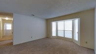 This single-floor space offers a clean, functional layout designed with comfort and practicality in mind. The main living area is a standout feature, characterized by its generous proportions and an abundance of natural light streaming in through a wide set of windows fitted with horizontal blinds. These windows create a bright, welcoming atmosphere while offering views of the outdoors. The walls are finished in soft, neutral beige tones that complement the plush wall-to-wall carpeting underfoot, contributing to a warm and understated aesthetic throughout. The ceiling features a textured popcorn finish, adding a classic touch to the interior. HVAC vents are thoughtfully integrated into both the ceiling and walls, ensuring efficient climate control throughout the space. The overall ambiance is calm and versatile, providing a blank canvas well-suited for a variety of furnishing styles and functional arrangements. The single-floor layout ensures seamless accessibility and easy navigation between areas, making it a practical and inviting environment.
