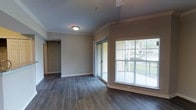This single-floor residence offers a clean, modern aesthetic defined by rich dark hardwood-style flooring that runs seamlessly throughout the open living area. Crown molding frames the neutral-toned walls, adding a refined architectural detail that enhances the overall sense of polish and quality. The main living space is bright and welcoming, benefiting from generous natural light that pours in through a large sliding glass door leading to a private balcony. The balcony features a railing and overlooks a lush, tree-lined outdoor area, providing a pleasant connection to the surrounding greenery. The open layout ensures a comfortable flow between the living area and the hallway leading to additional rooms, with interior doors finished in warm wood tones that complement the flooring. Recessed ceiling vents and flush-mounted ceiling lighting contribute to a clean and functional overhead design. The neutral warm-beige wall palette creates a versatile backdrop, making the space adaptable to a variety of interior design styles. Overall, the residence combines practical design with thoughtful finishes to deliver a comfortable and visually cohesive living environment.