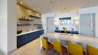 This contemporary single-story amenity space is designed with a cohesive modern aesthetic, combining bold accent colors, high-end finishes, and an open layout that encourages social interaction. The interior is anchored by a spacious kitchen area featuring a large granite-topped island with an undermount sink, navy blue cabinetry, open floating shelves with decorative accessories, and a suite of sleek pendant lights in a brushed metal finish. Vibrant mustard yellow mid-century modern bar chairs provide a striking contrast against the neutral tile flooring and crisp white walls.

The lounge area is defined by floor-to-ceiling grid-frame windows that flood the space with natural light and provide unobstructed views of a resort-style pool courtyard lined with palm trees and lounge seating. The seating arrangement includes a curved dark sectional sofa, modern swivel chairs in leather and patterned fabric, and patterned area rugs that add texture and warmth. A teal-accented ceiling and a statement sputnik chandelier add architectural interest overhead.

A niche alcove features a textured mosaic tile accent wall with integrated seating and yellow ceiling detail, serving as a focal design element. The overall ambiance is vibrant, polished, and resort-inspired throughout.