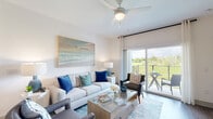 This single-floor residence is thoughtfully designed with a coastal aesthetic, blending bright, airy interiors with modern finishes throughout. The open-concept living room serves as the heart of the home, featuring light-toned walls, wood-look flooring, a ceiling fan, and large sliding glass doors that lead to a private balcony overlooking lush greenery — filling the space with abundant natural light. The kitchen is well-appointed with crisp white cabinetry, a textured tile backsplash, stainless steel appliances including a refrigerator with ice maker, electric range, and microwave. A peninsula with a light countertop and woven bar stools provides casual seating, while stylish industrial pendant lighting adds character overhead. The primary bedroom is generously sized with multiple large windows framing outdoor views, wood-look flooring, and ample natural light. A second bedroom echoes the same bright, welcoming atmosphere. Both bathrooms are finished with large-format marble-style tile flooring. The primary bath features a dual-sink vanity with white cabinetry, sleek black hardware, and illuminated mirrors. The second bath includes a shower-tub combination. A dedicated in-unit laundry area houses a full-size side-by-side washer and dryer with overhead shelving for added storage convenience.
