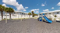 Located at 3640 Blue Fin Way in Fort Myers, Florida, this outdoor recreational area is a well-appointed community playground designed for active use and leisurely enjoyment. The space is fully enclosed by white metal fencing, providing a safe and defined boundary for the area. The ground is covered with a generous layer of rubber mulch, offering a cushioned, impact-absorbing surface throughout.

At the heart of the playground stands a colorful multi-component play structure featuring dual slides — one blue and one green — along with a climbing wall, making it an engaging centerpiece for children of various ages. Additional play equipment includes swings, spring riders, and teeter-totters, distributed across the space to accommodate multiple users simultaneously. A sturdy bench is positioned nearby, offering a comfortable spot for supervision or rest.

Tall, mature Florida palm trees are scattered throughout and around the perimeter, providing natural shade and lending a distinctly tropical aesthetic to the environment. The surrounding residential buildings are white with metal roofing and multi-pane windows, reflecting a clean, modern coastal architectural style. Under the open Florida sky, the area benefits from abundant natural light, creating a bright and inviting atmosphere. The combination of practical amenities and lush landscaping makes this a standout community recreational asset.