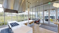 This contemporary single-floor space is defined by its striking floor-to-ceiling glass curtain walls, which flood the interior with natural light and offer sweeping views of the surrounding green landscape. The open-plan layout is anchored by a sleek demonstration kitchen outfitted with a professional-grade gas cooktop and a bold stainless steel range hood, paired with expansive white stone countertops that double as a casual dining and gathering surface. Colorful mixed seating — including mustard yellow and red chairs — adds a vibrant, welcoming contrast to the neutral palette of light wood ceiling panels and large-format stone tile flooring. A warm wood ceiling adds texture and warmth overhead, complementing the clean, minimalist aesthetic throughout. The dining and lounge area transitions effortlessly to an outdoor patio, visible through the glass walls and accessible via a full-length glass door, where additional seating and what appears to be an outdoor fireplace create an inviting extension of the interior. Recessed lighting and pendant fixtures provide layered illumination, enhancing the bright, airy ambiance. The overall design is polished, functional, and well-suited for communal gatherings or events.