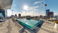 Located in the heart of Philadelphia at 1607 Sansom St, this exceptional rooftop amenity deck offers an elevated urban retreat with sweeping, unobstructed views of the city skyline, including iconic landmarks such as Liberty Place. The centerpiece of the space is a generously sized outdoor swimming pool with crystal-clear turquoise water, complemented by a separate hot tub — both finished with elegant mosaic tile detailing. Surrounding the pool, a broad stone-paved terrace provides ample room for lounging, furnished with sleek chaise lounge chairs and stylish seating arrangements. Large modern planters filled with lush greenery and seasonal flowers add a vibrant touch throughout the deck. A glass-enclosed indoor-outdoor lounge area runs along one side of the terrace, featuring floor-to-ceiling glass walls that frame the skyline views, high-top bar tables, and woven accent chairs — ideal for socializing or relaxing while sheltered from the elements. The open-air terrace wraps around the perimeter, offering multiple vantage points across the Philadelphia cityscape. The overall ambiance is sophisticated and resort-inspired, seamlessly blending modern design with the energy of downtown Philadelphia.