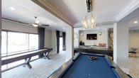 This single-floor entertainment space is thoughtfully designed to serve as a premium social and recreational hub. At the heart of the space sits a full-size pool table with a bold blue felt surface, positioned beneath an eye-catching cluster pendant chandelier featuring glass globe fixtures that add a sophisticated, artistic focal point to the room. Adjacent to the pool table, a sleek shuffleboard table occupies a carpeted area framed by dark drape panels and a ceiling fan, creating a distinct yet connected zone for gameplay. Natural light filters in through large windows and glass doors, enhancing the open, airy atmosphere. A built-in wet bar area lines one wall, complete with a mirrored backsplash, pendant lighting, and display accessories, making it ideal for hosting. A wall-mounted flat-screen television is centrally positioned above the bar counter, adding a media component to the entertainment mix. Decorative wall accents and recessed lighting throughout contribute to the polished, upscale ambiance. The combination of hard flooring and area rugs delineates each functional zone while maintaining a cohesive, contemporary aesthetic across the entire space.