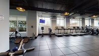 Located at 65 Bay St in Jersey City, NJ, this fitness center offers a professionally designed, single-floor workout facility equipped to support a wide range of training goals. The space features an industrial-modern aesthetic, with exposed ceilings finished in dark tones, rubber-tiled flooring, and a combination of pendant lighting and linear LED fixtures that create a focused, energetic atmosphere.

The cardio zone is generously outfitted with a row of treadmills, upright and recumbent stationary bikes, elliptical trainers, and stair climbers, many positioned along large windows that allow natural light to fill the space and provide views of the surrounding urban landscape. Mounted flat-screen televisions are positioned throughout for entertainment during workouts.

The strength and functional training area includes rowing machines, a cable and pulley multi-station unit, a power rack with TRX suspension system, plate-loaded machines, and various free-weight accessories. A glass-enclosed studio space is also present, offering a dedicated area for yoga, stretching, or group fitness activities, featuring wood-look flooring and bright, clean interiors. Full-length mirrors along the walls enhance the sense of openness throughout the facility, making it a comprehensive and well-appointed fitness destination.
