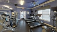 This single-level fitness center is thoughtfully designed to accommodate a wide range of workout styles within a spacious, open layout. The interior is finished with distinctive periwinkle-blue walls, crisp white crown molding, and dark rubber flooring, creating a cohesive and energizing environment throughout. Ceiling fans are positioned across the space to promote airflow and comfort during workouts.

The facility is divided into several functional zones. A dedicated cardio area features a row of treadmills and additional aerobic equipment positioned near windows with slatted blinds, allowing natural light to filter in. French doors in this section provide direct access to the outdoors. The strength training area is equipped with multiple Hoist cable machines and weight stations, supported by exercise instruction posters mounted on the walls for user reference.

A separate free weights zone includes a full dumbbell rack, stability balls, and a Smith machine for barbell training. Floor-to-ceiling mirrors line one wall of this area, enhancing the sense of space and allowing users to monitor their form. Fluorescent light fixtures supplement the natural light throughout, ensuring the entire facility remains well-lit. The layout provides ample floor space between equipment, supporting safe and comfortable movement throughout all areas.