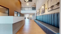 This contemporary single-floor clubhouse amenity space is designed with both elegance and functionality in mind. The grand entrance lobby immediately impresses with soaring double-height ceilings, a dramatic mezzanine level with metal railings, and a striking chandelier suspended overhead. Warm wood-look flooring flows throughout, complemented by a tufted teal banquette seating along a textured wall adorned with framed artwork and gold wall sconces, alongside a sleek reception desk.

Beyond the entry, a secondary reception area features warm wood-paneled walls and additional desk space, leading to various amenity zones. A comfortable lounge area offers plush caramel leather swivel chairs, a large flat-screen TV, built-in cabinetry, and rich teal accent walls. A private dining room provides an intimate setting with a solid wood table, upholstered chairs, bold patterned carpet, coffered ceiling detail, and curated wall art.

The well-appointed kitchen showcases light wood cabinetry, a decorative tile-accented island with bar seating, stainless steel appliances, and a coffee station. A casual social lounge with teal wainscoting opens to a media alcove with ample seating. The bright gathering hall, flooded with natural light through clerestory windows, features communal tables and direct sliding door access to an outdoor patio, seamlessly blending indoor and outdoor living.