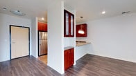 This single-floor residence offers a thoughtfully designed open-concept layout that seamlessly connects the kitchen and living areas. The kitchen is a standout feature, showcasing rich, warm-toned wood cabinetry with frosted glass upper cabinet inserts that add a refined touch. A peninsula with a stone countertop extends into the living space, providing both additional prep space and a casual dining option. Pendant lighting in a brushed metal finish hangs above the peninsula, casting a warm, inviting glow over the area. Stainless steel appliances, including a full-sized refrigerator, contribute a sleek, modern aesthetic that complements the cabinetry. White structural columns and walls create a crisp contrast against the deep wood tones, lending the space a clean, contemporary feel. Dark hardwood flooring runs throughout, adding warmth and continuity across the open floor plan. Recessed lighting is positioned strategically to ensure the space is well-lit, enhancing the overall bright and airy ambiance. The layout is practical and efficient, making excellent use of the available space while maintaining an attractive and welcoming atmosphere throughout.