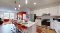 This thoughtfully designed single-floor residence combines modern finishes with bold, energetic design choices. The open-concept kitchen is a standout feature, showcasing white shaker cabinetry extending to the ceiling, stainless steel appliances including a full-size refrigerator and range, and a generous quartz-topped island with seating for multiple guests. Pendant lighting adds warmth overhead, while light hardwood flooring flows throughout the main living areas. Built-in shelving near the entry provides practical display and storage space.

The living room makes a striking impression with a vibrant accent wall, large windows that invite ample natural light, and views of the surrounding greenery. The space easily accommodates a full seating arrangement, creating a welcoming and lively atmosphere.

The bedrooms are generously proportioned, featuring plush carpeting and multiple windows that fill each room with natural light. The primary bathroom offers a floating white vanity with a large mirror and warm hardwood-style flooring, while a second full bathroom includes a tub-shower combination and clean, white finishes throughout.

Overall, this residence balances contemporary design with colorful personality, offering a comfortable and functional layout ideal for everyday living.