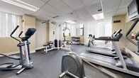 Located at 8 Silver Maple Crt in Brampton, Ontario, this fitness facility offers a well-organized, single-level layout designed to accommodate a variety of workout routines. The space features a suspended acoustic tile ceiling with recessed fluorescent lighting, and neutral beige walls that create a clean, functional environment. Large windows fitted with vertical blinds line multiple walls, allowing natural light to filter throughout the room while providing views of the surrounding outdoor area.

The facility is equipped with a comprehensive selection of exercise equipment, including multiple treadmills by Life Fitness, elliptical cross-trainers, and a stationary cycle. A multi-station cable machine supports a range of strength training exercises, while a dedicated free weights area features a tiered dumbbell rack with an assortment of weights and an adjustable workout bench. Protective floor matting is positioned in the stretching and free weight zones for safety and comfort.

A full-length wall mirror enhances the sense of space and supports form monitoring during workouts. A built-in cabinetry unit with a utility sink provides convenient access to water and storage. Informational fitness posters are displayed on the walls, adding a practical and motivational touch. The space is also equipped with a fire extinguisher and HVAC ventilation, ensuring safety and climate comfort throughout the facility.