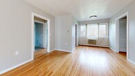 Located on Monroe Avenue in Rochester, NY, this 2-bedroom, 1-bathroom unit offers a practical and comfortable layout across a single floor. Warm-toned hardwood flooring flows consistently throughout the living areas and bedrooms, lending a cohesive and inviting feel to the space. The open living area is bathed in natural light from large windows dressed with vertical blinds, and includes a wall-mounted air conditioning unit and baseboard heating for year-round comfort. The spacious primary bedroom features windows overlooking the surrounding neighborhood, providing pleasant outdoor views and ample natural light. The second bedroom is a well-proportioned room with the same hardwood flooring and clean, neutral walls. The kitchen is a functional galley-style space equipped with warm wood cabinetry, light countertops, a stainless steel refrigerator, an electric range, and a double sink beneath a window with a view of greenery outside. The bathroom is neatly finished with marble-style tile walls, a soaking tub, a window with a curtain for natural light and privacy, and tile flooring. Throughout the unit, neutral gray walls and clean white trim create a bright, understated aesthetic that offers a versatile backdrop for any style.