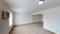 This single-floor residence features two bedrooms and one bathroom within a well-proportioned layout designed for efficient, comfortable living. The interior is finished with neutral tones throughout, creating a clean and versatile backdrop that suits a variety of décor styles.

The main living area is carpeted in a light beige tone that extends through the connected spaces, contributing to a cohesive, understated aesthetic. Flush-mount ceiling light fixtures provide consistent overhead illumination across the living and adjoining areas. Crisp white walls and classic wood baseboards frame each room, lending a tidy, well-maintained appearance.

An open passage connects the primary living space to an adjacent area, enhancing the sense of flow and openness within the home. The layout appears straightforward and practical, supporting easy navigation between rooms on the single floor. Electrical outlets are conveniently positioned along the walls, and a thermostat is visible, indicating climate control capabilities.

Overall, this residence presents a simple, move-in-ready environment with a neutral palette, functional lighting, and a connected layout that maximizes the usability of its comfortable square footage.