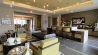This elegant single-floor space showcases a modern, contemporary design that seamlessly blends comfort with sophistication. The main lounge area is anchored by a coffered ceiling with warm ambient uplighting and a series of globe pendant lights that cast a welcoming glow throughout the space. Large-format gray tile flooring grounds the interior and provides a sleek, neutral foundation for the carefully curated furnishings.

Seating is arranged thoughtfully across the lounge, featuring chartreuse green upholstered chairs and a striking tufted dark sofa accented with bold black-and-white patterned pillows, creating a dynamic contrast of color and texture. A built-in bar or counter area lines one wall, complete with backlit stone tile cladding, mounted flat-screen television, and modern bar seating, making it well-suited for social gatherings or casual meetings.

Decorative elements throughout include fresh floral arrangements, framed artwork, and sculptural accents that add personality and life to the environment. An adjacent room, visible through wide doorways, continues the refined aesthetic with additional seating and abundant natural light. The overall ambiance is polished and contemporary, offering a space that is both functional and visually impressive.