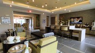 This elegant single-floor space showcases a modern, contemporary design that seamlessly blends comfort with sophistication. The main lounge area is anchored by a coffered ceiling with warm ambient uplighting and a series of globe pendant lights that cast a welcoming glow throughout the space. Large-format gray tile flooring grounds the interior and provides a sleek, neutral foundation for the carefully curated furnishings.

Seating is arranged thoughtfully across the lounge, featuring chartreuse green upholstered chairs and a striking tufted dark sofa accented with bold black-and-white patterned pillows, creating a dynamic contrast of color and texture. A built-in bar or counter area lines one wall, complete with backlit stone tile cladding, mounted flat-screen television, and modern bar seating, making it well-suited for social gatherings or casual meetings.

Decorative elements throughout include fresh floral arrangements, framed artwork, and sculptural accents that add personality and life to the environment. An adjacent room, visible through wide doorways, continues the refined aesthetic with additional seating and abundant natural light. The overall ambiance is polished and contemporary, offering a space that is both functional and visually impressive.