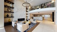 This elegant lobby presents a striking first impression through its double-height ceilings and refined contemporary design. The centerpiece is a dramatic floor-to-ceiling marble accent wall framing a sleek linear fireplace, flanked by dark floating shelves displaying carefully curated decorative objects. A bold circular chandelier anchors the seating area below, where neutral-toned sofas and chairs are arranged around metallic gold and black accent tables atop an abstract blue-and-grey area rug.

The reception desk, clad in veined stone, sits prominently within the space, complemented by warm pendant lighting and an illuminated shelving backdrop. A glass-railed mezzanine level overlooks the main floor, adding architectural depth and visual interest.

The entry foyer introduces the space with large floor-to-ceiling windows that flood the area with natural light, alongside a sculptural art installation and built-in display shelving. An adjacent secondary lounge features dark leather seating, soft drapery, and high ceilings, offering a quieter retreat.

Throughout, large-format porcelain tile flooring, warm wood accents, curated artwork, and carefully placed sculpture contribute to a cohesive, upscale ambiance that is both welcoming and architecturally refined.