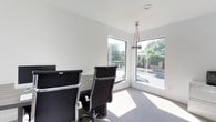 This single-floor office space presents a clean, contemporary design with a strong emphasis on minimalism and functionality. The interior is finished in crisp white walls and neutral-toned carpeting, creating a bright and professional atmosphere throughout.

The workspace features a dedicated office area outfitted with a modern desk and ergonomic high-back chairs with chrome accents, ideal for focused work or small meetings. A standout design element is the contemporary pendant light fixture with distinctive red cord detailing, suspended from the ceiling to add a subtle artistic touch to the otherwise understated décor.

Large corner windows are a defining architectural feature, allowing generous amounts of natural light to flood the interior while offering open views of the surrounding landscaped outdoors, including mature trees and street scenery. This combination of light and greenery contributes to a refreshing and comfortable work environment.

Recessed ceiling lighting complements the natural light, ensuring the space remains well-illuminated throughout the day. The overall layout is efficiently arranged, making practical use of the available floor area while maintaining an uncluttered and orderly feel. This office space is well-suited for professionals seeking a modern, light-filled environment that balances style with practicality.