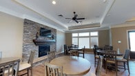 This single-floor common gathering space is thoughtfully designed to balance comfort and functionality. The room's most prominent feature is a floor-to-ceiling stacked stone fireplace wall, which anchors the space with a warm, rustic charm. A dark wood mantel shelf adds character, while a wall-mounted flat-screen television above makes the area suitable for both relaxed viewing and social gatherings.

The elevated tray ceiling with crown molding lends an architectural refinement to the room, complemented by a centrally positioned ceiling fan that promotes air circulation. Recessed lighting throughout ensures the space is well-lit at all times.

Multiple round dining tables with wooden chairs are arranged throughout, providing flexible seating for groups of various sizes. The light hardwood flooring ties the neutral, warm-toned walls together cohesively. Natural light floods the room through a large multi-pane window fitted with horizontal blinds, brightening the dining area and creating an inviting atmosphere. The overall design strikes a balance between casual comfort and polished detail, making it well-suited as a communal lounge or dining area.