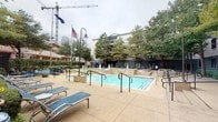 This outdoor amenity space is centered around a generously sized resort-style swimming pool with crystal-clear blue water and multiple entry points via metal handrail staircases. A standout feature is the elevated fountain structure positioned at the heart of the pool area, with dual water jets that create a dynamic, resort-like ambiance. The pool deck is finished in a warm-toned concrete surface and is outfitted with numerous blue sling-style lounge chairs and chaise lounges, offering ample space for relaxation and sunbathing.

The courtyard is framed by multi-story buildings with classic architectural detailing, including arched colonnades, balconies, and stucco facades, lending the space an upscale, community-centered character. Mature trees are distributed throughout the area, providing natural shade and greenery that soften the hardscape. American and state flags are prominently displayed on flagpoles near the pool.

A secondary outdoor lounge area adjacent to the pool features bar-height seating and tables on a wide concrete patio, ideal for social gatherings. Access to the space is controlled through ornamental wrought-iron security gates with electronic access hardware. Decorative ceramic planters with seasonal flowers add color and charm to the entryways, enhancing the welcoming atmosphere of this well-appointed outdoor retreat.