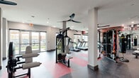 Located at 269 S State Highway 360 in Mansfield, Texas, this well-appointed fitness center offers a comprehensive workout environment designed for a wide range of exercise routines. The single-floor facility is organized into distinct zones, each tailored to specific training needs.

The cardio area is lined with a row of treadmills and a stair climber, positioned alongside large windows and glass doors that invite ample natural light and provide views of the surrounding outdoor greenery. Overhead ceiling fans keep the space comfortable during high-intensity sessions.

A dedicated strength training zone houses a multi-function cable machine, a full rack of dumbbells, and a robust multi-station weight machine, all set against a bold terracotta accent wall adorned with motivational fitness imagery and a wall-mounted flat-screen TV.

A separate studio space provides room for cycling classes, equipped with stationary bikes, an additional flat-screen display, and a colorful geometric floor design in orange and gray. Glass partition walls connect the studio to the main gym floor, maintaining visual openness throughout.

The flooring throughout the facility features durable rubberized surfaces in coordinating red, gray, and dark charcoal tones, reinforcing the energetic and modern aesthetic. Recessed lighting and multiple large windows ensure the space remains bright and inviting at all times.