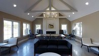 This single-story space is defined by its striking vaulted ceiling, which rises dramatically with exposed white-painted beams forming a classic cathedral structure. A statement brass chandelier with multiple exposed bulbs hangs at the apex, providing warm ambient lighting that complements the recessed lighting throughout the space.

The room is anchored by a clean, white-paneled fireplace centered on the main wall, above which hangs a nature-inspired framed artwork, adding a touch of character. The warm taupe walls contrast with crisp white trim and ceiling elements, lending the space a neutral, sophisticated palette.

Polished concrete floors in a cool grey tone run throughout, offering a modern counterpoint to the traditional architectural details. Multiple white-framed windows line the walls, allowing natural light to filter in from multiple directions and creating an airy, open atmosphere.

The furnishings are thoughtfully arranged, combining dark upholstered seating and task-oriented workstations along the perimeter with a comfortable sofa grouping in the foreground. This layout supports both collaborative work and relaxed conversation, making the space highly functional and versatile.