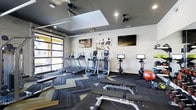 This single-level fitness center offers a thoughtfully designed workout space with a contemporary aesthetic. The room features a striking herringbone-patterned carpet in dark tones with gold accents, complemented by a dark ceiling with recessed lighting and a central skylight that allows natural light to filter in. A large glass garage-style door along one wall provides additional natural light and views of the exterior, creating an open, airy atmosphere.

The cardio zone is well-stocked with multiple treadmills and elliptical machines, each equipped with individual digital screens for entertainment or workout tracking. Stationary and Peloton-style cycling bikes are also available for low-impact training. Two wall-mounted flat-screen televisions are positioned for easy viewing throughout the space, alongside motivational artwork depicting fitness themes.

A dedicated free-weight and functional training area includes weight benches and exercise balls, offering versatility for strength training routines. The overall design balances modern industrial elements with practical functionality, creating an inviting and energizing environment suitable for a wide range of fitness activities.