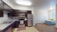 This single-story unit is designed with a functional, modern layout that integrates the kitchen, living, and sleeping areas into a cohesive open-concept floor plan. The kitchen serves as a focal point, outfitted with a full suite of stainless steel appliances including a refrigerator, electric range with over-the-range microwave, and a dishwasher. Rich, dark espresso-toned cabinetry extends to the ceiling, providing ample storage, while granite-style countertops and a textured tile backsplash add a refined aesthetic. Light wood-look flooring runs through the kitchen area, transitioning to carpeting in the living and sleeping zones. The living area is bright and simply appointed, featuring a window that admits natural light and accommodates comfortable seating. The sleeping area, visible in the aerial view, offers adequate space for a standard bed and additional furnishings. The bathroom is clean and well-appointed, featuring a granite-topped vanity with dark cabinetry, a large mirror, and tile flooring. Throughout the unit, neutral wall tones and consistent finishes contribute to a cohesive, understated atmosphere well-suited for everyday living.