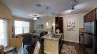 This single-floor residence showcases an open-concept layout that seamlessly integrates the kitchen and living area into one cohesive, functional space. The kitchen serves as a central hub, featuring a peninsula island with granite countertops and a double basin sink, dark espresso-toned cabinetry reaching toward the ceiling for ample storage, and elegant pendant lighting hanging overhead. Track lighting along the ceiling adds targeted illumination, complementing the warm, neutral tones of the walls. Rich, dark hardwood flooring runs throughout, grounding the space with a sense of warmth and continuity. A decorative patterned area rug near the kitchen adds a pop of color and texture. The living area beyond is generously sized, anchored by a ceiling fan with light fixture and bathed in natural light from a large window with horizontal blinds. Comfortable seating arranged around a central area creates an inviting atmosphere, while a coastal-inspired wall painting adds a relaxed, artistic touch. Decorative wall art in the kitchen area further enhances the curated aesthetic. The overall ambiance is modern yet warm, combining practical design with thoughtful decorative details throughout.