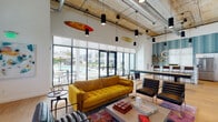This single-floor community space showcases an impressive industrial-chic design, highlighted by soaring exposed concrete ceilings with visible ductwork and a striking array of pendant and track lighting. The open-plan layout flows seamlessly from a comfortable lounge area to a fully equipped kitchen, creating a versatile, social environment throughout.

The lounge area is anchored by a bold mustard-yellow tufted sofa paired with sleek black leather chairs, all set atop a vibrant, multicolored area rug over warm hardwood flooring. A wall-mounted flat-screen TV and floating wood shelving complete the media corner. Whimsical surfboard décor suspended from the ceiling adds a distinctive coastal character to the space.

Floor-to-ceiling glass doors and windows flood the interior with abundant natural light and offer direct views of an outdoor pool and lounge area, reinforcing the resort-like atmosphere. High clerestory windows along additional walls further enhance the bright, airy feel.

The kitchen is a standout feature, finished with crisp white cabinetry, light quartz countertops, and a bold teal brick-pattern tile accent wall. Open wooden shelving, circular halo pendant lights, and stainless steel appliances—including a French door refrigerator—lend both style and functionality. A generously sized island with bar seating makes this kitchen ideal for casual gatherings and community events.