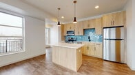 This single-floor space showcases a thoughtfully designed open-concept layout with consistent wood-look flooring that flows through the main living and kitchen areas. The kitchen serves as a focal point, featuring light wood flat-front cabinetry accented by matte black hardware, a striking teal hexagonal ceramic tile backsplash, and a full suite of stainless steel appliances including a refrigerator, range, microwave, and dishwasher. A central island with a white quartz-style countertop provides additional workspace and is complemented by two pendant lights with dark cone-shaped shades. A deep undermount sink with a matte black faucet adds a refined touch.

The living area is bright and open, benefiting from large windows that invite generous natural light and offer outdoor views, including a glimpse of an exterior balcony railing. The bedroom is a calm, neutral space finished with plush carpet flooring and a sizeable window providing ample daylight. The bathroom is clean and functional, featuring a wood-toned vanity with white countertop, matte black hardware, a soaking tub, and tile flooring. Throughout, the design maintains a cohesive modern aesthetic with a neutral, warm palette.