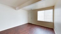 This two-floor space presents a clean, move-in-ready aesthetic with thoughtful design details throughout. The main living area, visible in the image, showcases rich, dark hardwood-style flooring that runs continuously across the room, creating a warm and grounded atmosphere. Neutral beige and white walls provide a versatile backdrop, making the space easy to personalize. A structural beam accent on the ceiling adds a subtle architectural element while lending a sense of definition to the room. Natural light filters in through a window fitted with horizontal blinds, offering both brightness and privacy control. A paneled interior door provides access to an adjacent room or storage area, contributing to the overall functional flow of the layout. The space spans two floors, suggesting additional living, sleeping, or utility areas beyond what is immediately visible. The overall ambiance is understated and practical, with a neutral palette and durable finishes that suit a variety of uses. The combination of natural light, quality flooring, and clean walls gives the space an open and welcoming feel throughout.
