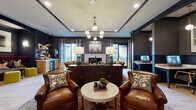 Located at 1145 Chambers Rd in Columbus, Ohio, this elegantly designed single-floor communal lounge space offers a richly layered interior that balances comfort with refined style. The main gathering area is anchored by a marble-surround fireplace and features deep, dark-painted walls that create an intimate and sophisticated ambiance. Plush leather seating, a large sectional sofa, and ornate area rugs contribute to a warm, club-like atmosphere, while a multi-globe brass chandelier serves as a striking focal point overhead. A built-in wet bar and kitchenette area provide convenient refreshment options, complete with open shelving and under-counter refrigeration. An adjacent nook offers a cozy banquette seating area upholstered in classic plaid, paired with velvet accent chairs and wall sconces, ideal for smaller gatherings or quiet retreats. The reception area features a sculptural white desk set against bold blue cabinetry and a patterned tile accent wall, adding a vibrant, artistic contrast. Light hardwood herringbone flooring flows throughout, complemented by layered vintage-style rugs. Sliding glass doors lead to an outdoor deck, filling the space with natural light and providing seamless indoor-outdoor connectivity.