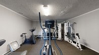 Located at 6016 Sunrise Vista Dr in Citrus Heights, CA, this dedicated fitness room offers a well-organized workout space designed to support a range of exercise routines. The room features a textured ceiling with crown molding detail and neutral gray walls throughout, creating a clean, functional environment. Carpet flooring covers the entire space, providing a firm yet comfortable surface underfoot.

The room is equipped with an impressive variety of fitness equipment. Cardio options include treadmills and an elliptical trainer, while a stationary spin bike offers additional variety. Strength training is well represented by a Hoist multi-station cable machine and a vertical knee raise/dip station. A dedicated stretching bench and free weights round out the available equipment.

A large wall-mounted mirror spans one side of the room, visually expanding the space and allowing users to monitor their form during workouts. Overhead flush-mount lighting illuminates the room evenly. A glass-paned door provides access to an adjacent common area, allowing natural light to filter in and offering a sense of openness. A portable fan is also on hand for added comfort during workouts. Compact yet thoughtfully equipped, this fitness room provides residents with convenient access to a comprehensive exercise facility.