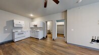 Located at Graystone Landing on East Berry Street in Fort Wayne, Indiana, this single-floor unit offers a practical and well-organized layout with a clean, modern aesthetic. Warm wood-style luxury vinyl flooring runs consistently throughout the space, complemented by neutral gray walls and gray baseboards that create a cohesive, contemporary feel.

The kitchen is equipped with white cabinetry, light stone-style countertops, a double-basin stainless steel sink, a dishwasher, an electric range with overhead microwave, and a full-size refrigerator. A bold blue entry door adds a distinctive accent near the kitchen area. The layout allows the kitchen to flow naturally toward the main living space, which features a ceiling fan and a wall-mounted AC unit beneath a window that brings in natural light.

Additional rooms include a bedroom with a ceiling fan, a window, a wall-mounted AC unit, and closet access. A secondary room provides flexible use and also connects to the main hallway. The bathroom is well-appointed with a walk-in corner shower with chrome framing, a single-sink vanity with white countertop, a large mirror, and an in-unit washer, adding everyday convenience to the space.
