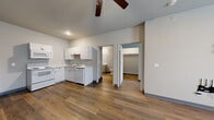 Located at Graystone Landing on East Berry Street in Fort Wayne, Indiana, this single-floor unit offers a practical and well-organized layout with a clean, modern aesthetic. Warm wood-style luxury vinyl flooring runs consistently throughout the space, complemented by neutral gray walls and gray baseboards that create a cohesive, contemporary feel.

The kitchen is equipped with white cabinetry, light stone-style countertops, a double-basin stainless steel sink, a dishwasher, an electric range with overhead microwave, and a full-size refrigerator. A bold blue entry door adds a distinctive accent near the kitchen area. The layout allows the kitchen to flow naturally toward the main living space, which features a ceiling fan and a wall-mounted AC unit beneath a window that brings in natural light.

Additional rooms include a bedroom with a ceiling fan, a window, a wall-mounted AC unit, and closet access. A secondary room provides flexible use and also connects to the main hallway. The bathroom is well-appointed with a walk-in corner shower with chrome framing, a single-sink vanity with white countertop, a large mirror, and an in-unit washer, adding everyday convenience to the space.