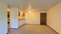 This single-floor unit offers a straightforward, functional layout centered around an open living area. The space is finished with wall-to-wall carpet flooring in a neutral tone, contributing to a warm and understated ambiance throughout. A ceiling fan with an integrated light fixture is centrally mounted, providing both air circulation and general illumination to the room.

One of the defining features of the layout is the partial wall with a pass-through counter that separates the living area from the kitchen. This design element maintains an open feel while defining each zone. Above the counter, medium-toned wood cabinetry provides storage and adds a touch of warmth to the otherwise light, neutral color palette of the walls and ceiling.

The ceiling features a subtle textured finish, and trim baseboards run along the perimeter of the room, offering clean architectural detail. A solid wood-toned entry door with a deadbolt provides the main point of access to the unit, positioned along one wall of the living area. The overall ambiance is clean and practical, with a simple design suited for comfortable everyday living.