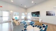 Located at 8855 McGallion Rd in Houston, TX, this single-floor community lounge offers a well-appointed, open living area designed for comfort and social interaction. The space features a contemporary aesthetic anchored by crisp white walls, warm light wood flooring, and elevated ceilings that contribute to an airy, spacious ambiance.

The living room is the heart of the space, divided into distinct seating zones united by bold blue, white, and tan geometric area rugs. One zone centers around a wall-mounted flat-screen TV and a light wood console table, flanked by modern lounge chairs and round white accent tables. A second seating area offers a tan sofa, upholstered armchairs in soft blue, and additional ottomans, creating an inviting gathering space. Eye-catching abstract wall art in blue and gray tones adds a creative focal point.

Large windows fitted with roller shades line the walls, flooding the interior with natural light and offering views of the surrounding outdoor area. A set of full-length glass double doors with transom panels opens to the exterior, seamlessly connecting the indoor and outdoor environments. Recessed and pendant lighting provide warm, layered illumination throughout. The overall design balances modern style with functional, welcoming comfort.