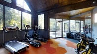 This fitness center offers a thoughtfully designed workout environment that seamlessly blends functionality with natural surroundings. The space features a warm, wood-paneled ceiling that adds a rustic yet refined architectural character, complemented by dark-framed walls that create a bold, modern contrast.

Large windows and glass-panel doors line multiple walls, flooding the interior with abundant natural light and providing expansive views of the surrounding greenery. This connection to the outdoors creates an energizing and refreshing atmosphere for exercise.

The flooring is a standout design element, featuring a striking geometric pattern in bold shades of orange, red, and white, adding a dynamic visual appeal to the space. The fitness equipment includes multiple elliptical and cardio machines positioned to take advantage of the natural light and views. A dedicated free weights area with a full dumbbell rack is situated along the far wall, catering to strength training needs.

Overall, the space strikes a balance between an inviting, nature-inspired aesthetic and a well-equipped, practical fitness environment suitable for a variety of workout routines.