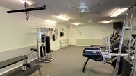 The fitness center at Sugar Mill Creek is a single-floor exercise facility designed to support a variety of workout routines. The room features a neutral color palette with light green accent walls and white trim, creating a clean and functional environment. Ceiling fans provide consistent air circulation throughout the space, complementing the fluorescent lighting that ensures the area is well-lit during use. The flooring consists of durable carpeting suited for a gym setting. Equipment includes a multi-function cable/weight machine and additional strength training apparatus, offering options for both resistance and functional training. A wall-mounted clock keeps exercisers on schedule, and a folding chair is available for rest or stretching. Mirrored wall panels on one side enhance the sense of openness while allowing users to monitor their form during workouts. A paneled door at the rear provides access to additional areas of the facility. The overall layout is open and uncluttered, allowing comfortable movement between equipment stations.