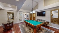 Located at 4401 E 46th St in North Little Rock, AR, this dedicated game room is a well-appointed entertainment space that blends classic elegance with leisure-focused design. The room is anchored by a full-size pool table with a green felt surface, positioned atop a decorative area rug that complements the rich, warm-toned hardwood floors throughout. A chain-hung billiard light fixture provides focused illumination over the table, while vaulted ceilings add a sense of vertical openness and grandeur to the space. A standout architectural feature is the marble-surround fireplace set against a stone-clad accent wall, flanked by a cozy seating area with leather armchairs and a bistro-style table — ideal for relaxed conversation. Ornate gold-framed mirrors and artwork adorn the walls, lending the room a refined, classical aesthetic. A wall-mounted flat-screen television is positioned for easy viewing, accompanied by an elegantly carved bench and decorative curio cabinets with gold detailing. French doors with glass panes connect the game room to adjacent areas of the property, allowing natural light to filter in. The overall ambiance is one of sophisticated leisure, combining entertainment functionality with upscale interior design sensibilities.