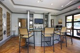This well-appointed single-floor community lounge space combines comfort and style in a cohesive, inviting design. The main seating area features a wraparound upholstered sectional sofa adorned with striped accent pillows, positioned beneath a tray ceiling with crown molding. Framed landscape artwork and textured woven wall panels paired with decorative silver chargers add depth and character to the walls, creating a warm, curated ambiance. Roman shades on the windows allow natural light to filter softly into the room. The adjacent area includes a built-in wet bar with dark countertops, glass-front cabinetry, a wall-mounted flat-screen television flanked by open shelving, and a compact refrigerator — ideal for entertaining. French doors connect the interior to a secondary lounge zone, which flows into a dining area with a round glass-top table and woven cane chairs. Large floor-to-ceiling glass doors open directly to an outdoor pool deck, offering abundant natural light and seamless indoor-outdoor connectivity. The polished concrete flooring in rich terra cotta tones ties all areas together, complemented throughout by soft grey walls and dark wood trim accents that lend the space a refined, resort-inspired atmosphere.