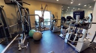 This fitness center offers a comprehensive workout environment designed to support a wide range of exercise routines. The space is outfitted with an array of cardio machines, including elliptical trainers, positioned along one side of the room for easy access. A multi-function cable weight machine anchors the strength training area, providing users with versatile resistance training options. Exercise balls in varying sizes are also available, adding flexibility for core and balance workouts. The dark rubber flooring provides a durable, slip-resistant surface well-suited for high-activity use. A large mirror spans one full wall, enhancing the sense of openness and allowing users to monitor their form during exercise. Natural light filters in through a window overlooking lush greenery, creating a bright and energizing atmosphere. The warm-toned walls contribute to an inviting ambiance, while recessed ceiling lighting ensures the space remains well-lit throughout. Informational notices are posted on the walls, offering guidance for users. The overall layout is thoughtfully organized to maximize the available floor space, making this fitness center a practical and fully functional amenity.