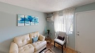 This single-floor unit is characterized by a cohesive soft blue-gray color palette carried throughout each room, creating a calm and unified atmosphere. Textured ceilings and wood-style flooring are consistent features that add warmth and character to the space.

The living area offers natural light through a large window dressed with sheer lace curtains, complemented by a wall-mounted air conditioning unit. The room accommodates comfortable seating and decorative wall art, lending a relaxed yet styled ambiance.

The kitchen is efficiently designed with white cabinetry lining the walls, providing ample storage. White appliances — including a range, dishwasher, and refrigerator — maintain a clean, cohesive look. A compact dining area with a white table and chairs sits adjacent, illuminated by overhead fluorescent lighting.

The bedroom features multiple windows with sheer curtains that allow generous natural light to filter in. A wall-mounted air conditioning unit, a full-length mirror, and decorative canvas artwork above the bed contribute to the room's bright and organized feel. Indoor greenery adds a fresh, natural touch.

The bathroom includes a tiled tub-shower combination finished in neutral tones, accented by colorful décor that adds a lively contrast to the otherwise serene color scheme throughout the unit.